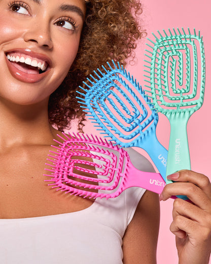 Smiling woman with curly hair holds three colorful UNbrush hairbrushes—pink, blue, and teal—against a pink background.