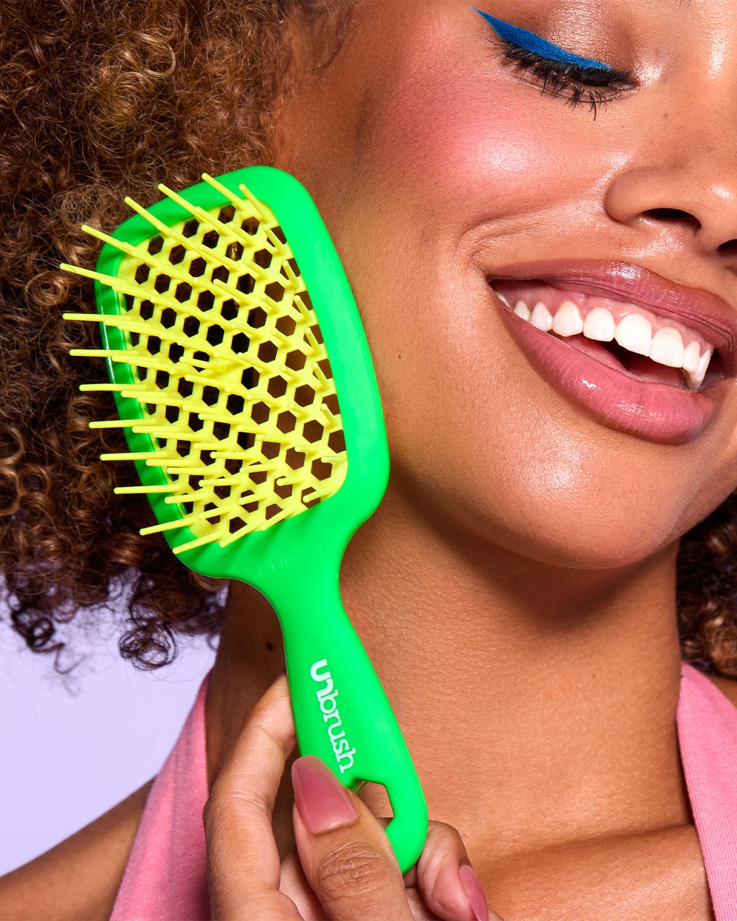 Smiling woman with curly hair holds a green and yellow detangling UNbrush Mini, highlighting its use for styling textured or curly hair.