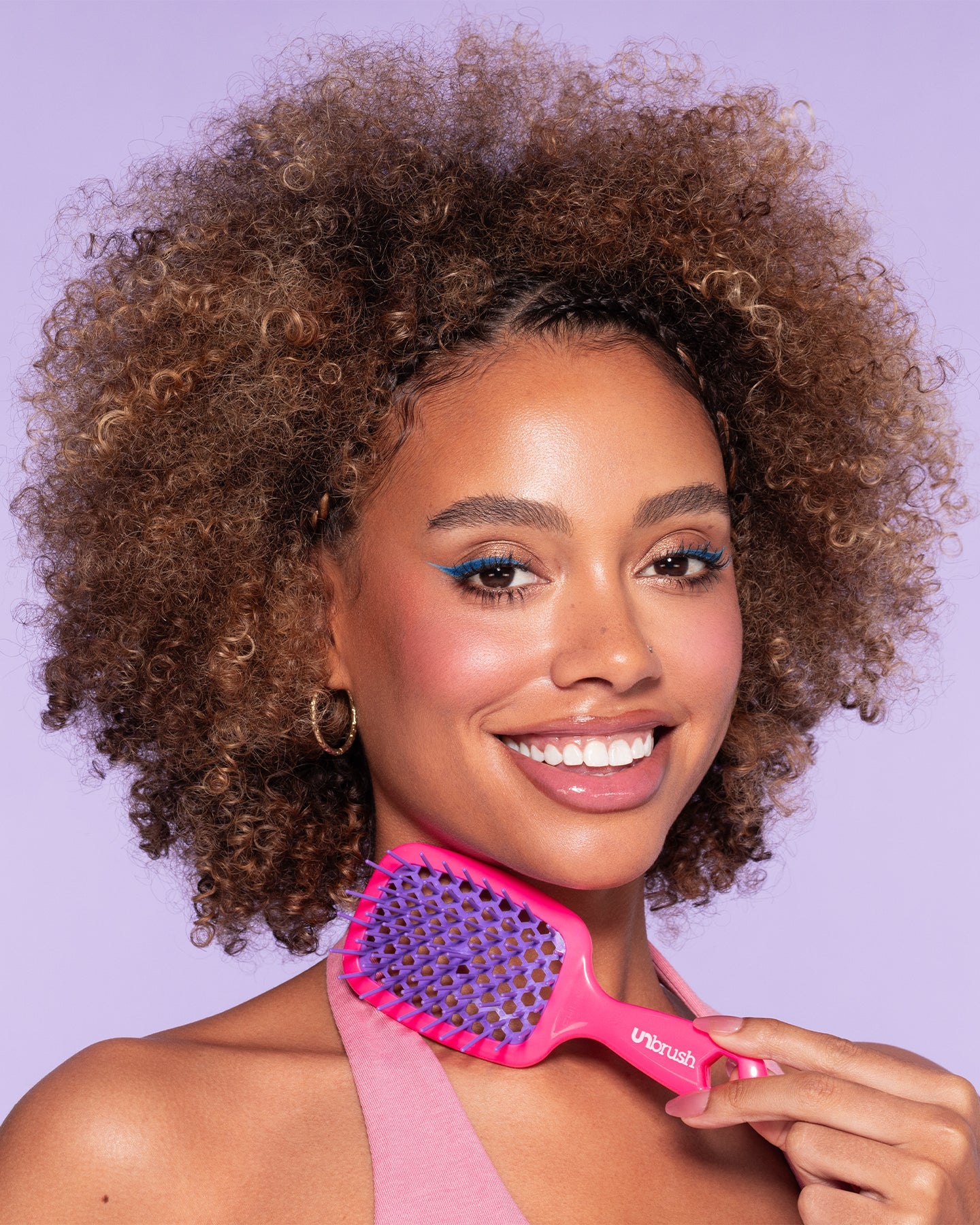 Smiling woman with curly hair holds a pink and purple detangling UNbrush Mini, highlighting its use for styling textured or curly hair.
